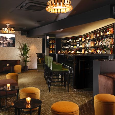 โรงแรมเซนต์ มาร์ติน เลน ลอนดอน ภายนอก รูปภาพ The photo depicts a stylish bar or lounge interior. The space features a dark, sophisticated color scheme with plush seating, including brown couches and vibrant yellow stools. A illuminated bar area is prominently displayed, lined with various bottl
