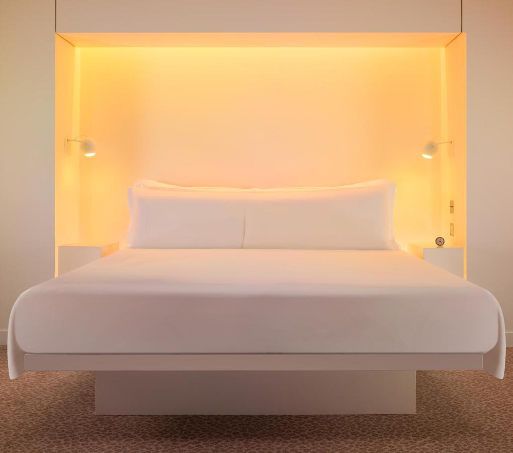 โรงแรมเซนต์ มาร์ติน เลน ลอนดอน ภายนอก รูปภาพ The photo shows a modern and minimalist bedroom setup featuring a large white bed with two pillows. The bed is positioned against a glowing orange backdrop, which adds warmth to the ambiance. On either side of the bed, there are small nightstands equ