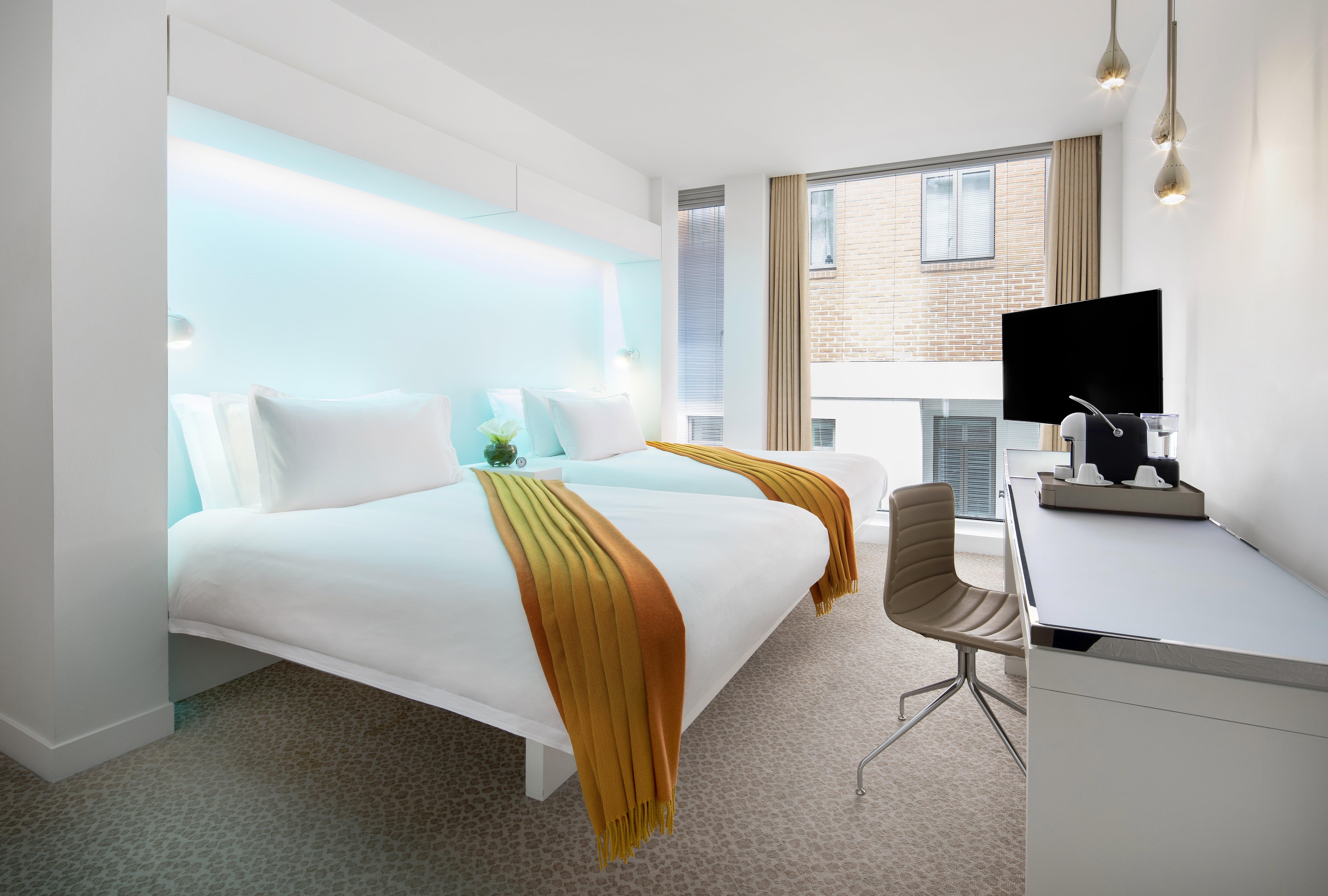 โรงแรมเซนต์ มาร์ติน เลน ลอนดอน ภายนอก รูปภาพ The photo shows a modern hotel room with a clean, contemporary design. There are two beds, each with white bedding and vibrant orange and yellow throws. Behind the beds, there is a glowing blue accent wall, adding a stylish touch. The room features a