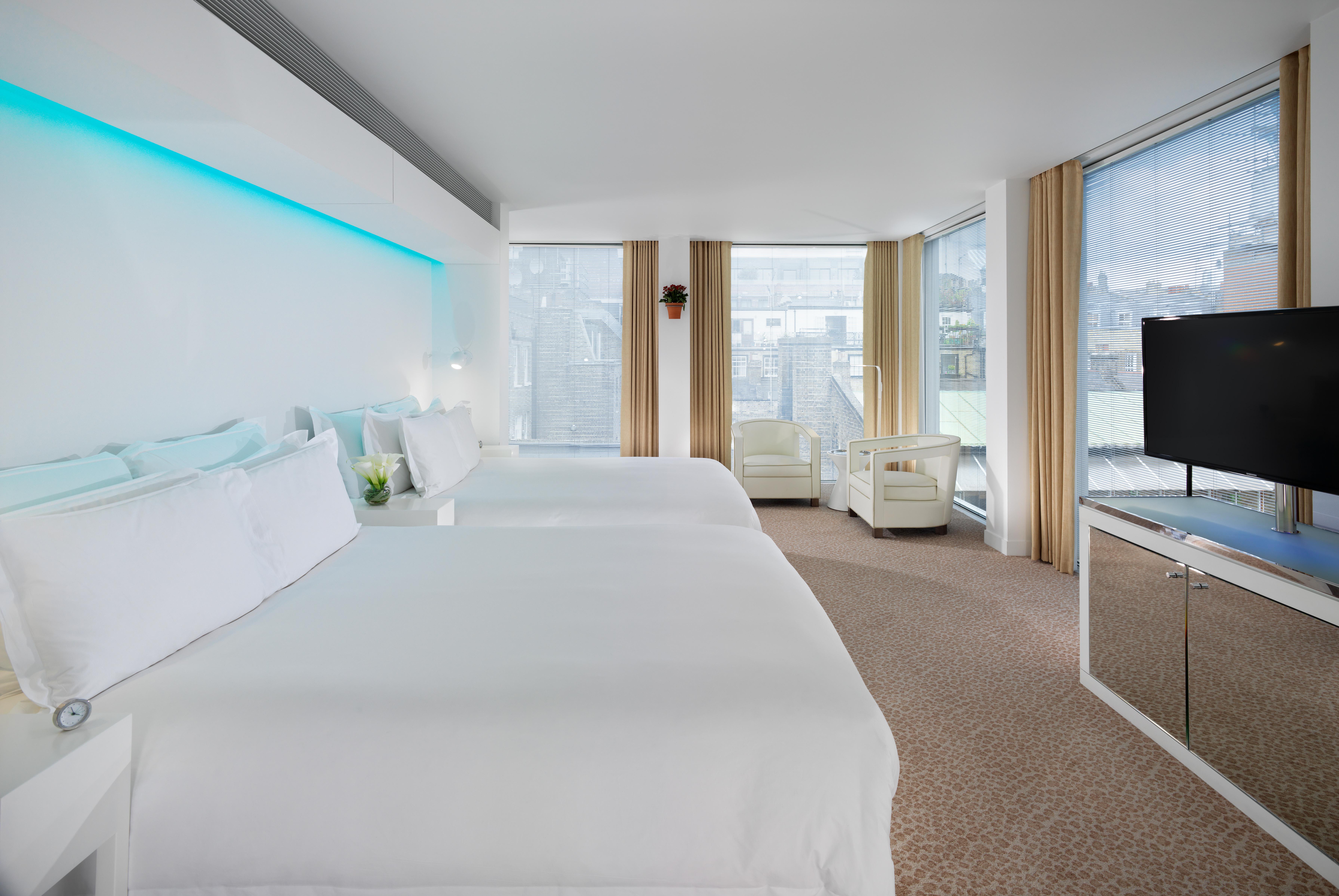 โรงแรมเซนต์ มาร์ติน เลน ลอนดอน ภายนอก รูปภาพ The photo shows a modern hotel room featuring two large beds with white bedding. The room has large windows with light-colored curtains, allowing natural light to fill the space. There is a small seating area with a white sofa and a bedside table. A 