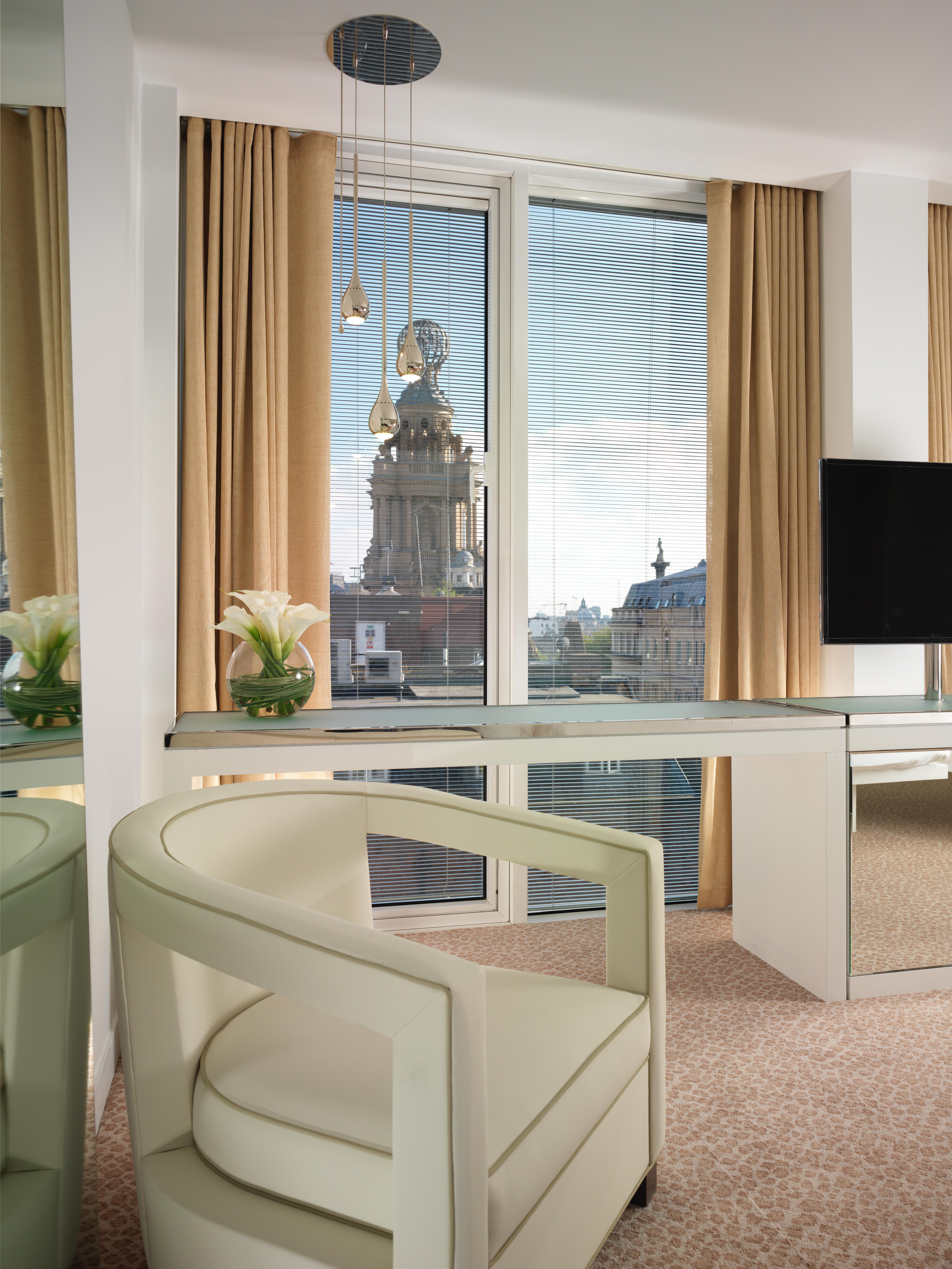 โรงแรมเซนต์ มาร์ติน เลน ลอนดอน ภายนอก รูปภาพ The photo shows a modern interior space, possibly a hotel room or office, featuring a sleek glass desk and a contemporary chair. There’s a vase of white flowers on the desk, and large windows draped with beige curtains provide a view of a building ou