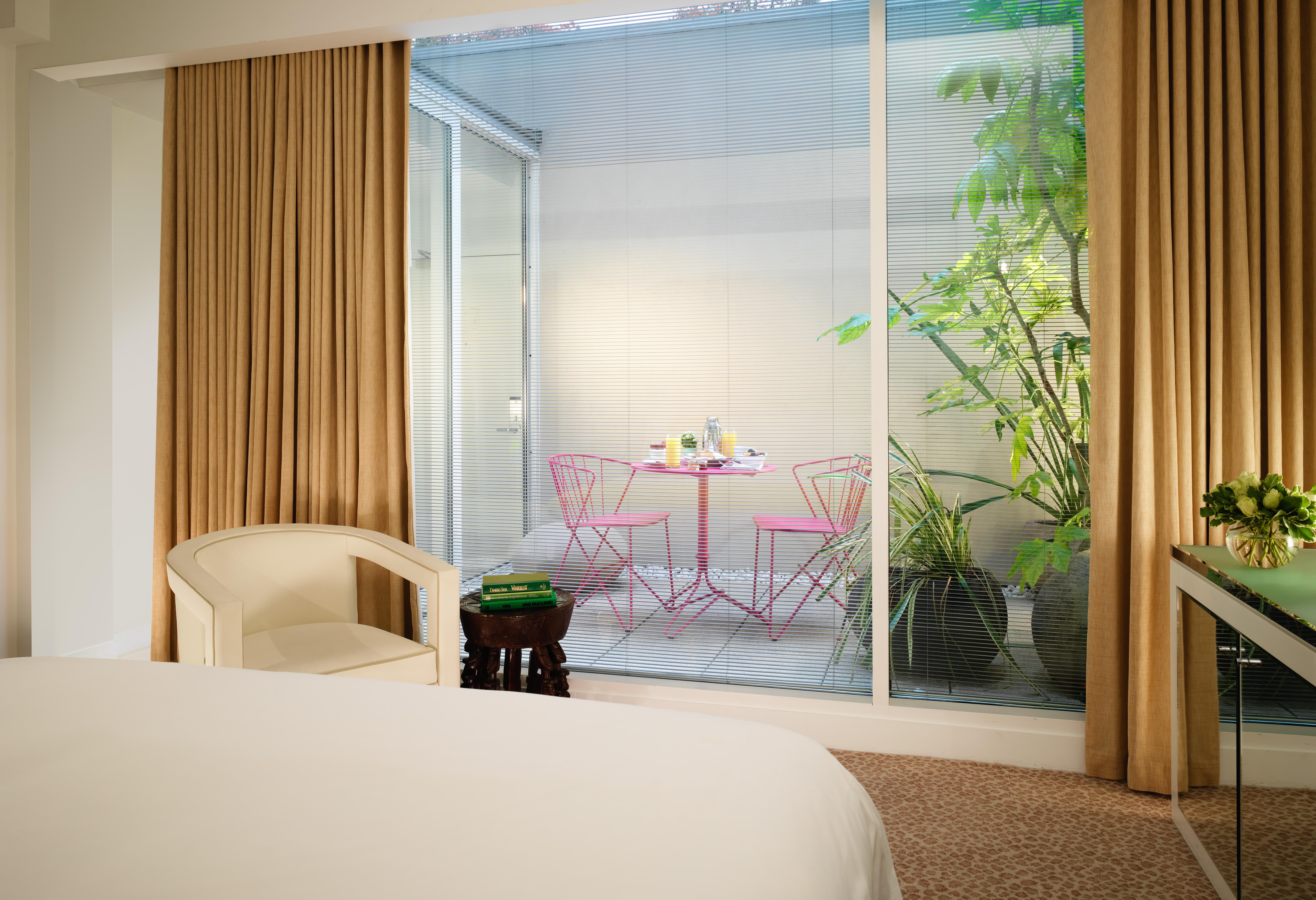 โรงแรมเซนต์ มาร์ติน เลน ลอนดอน ภายนอก รูปภาพ The photo depicts a modern room with a bed visible in the foreground. Large glass windows allow natural light to enter, showcasing a small outdoor space. Beyond the glass, there is a charming patio area featuring a round table and two pink chairs, su