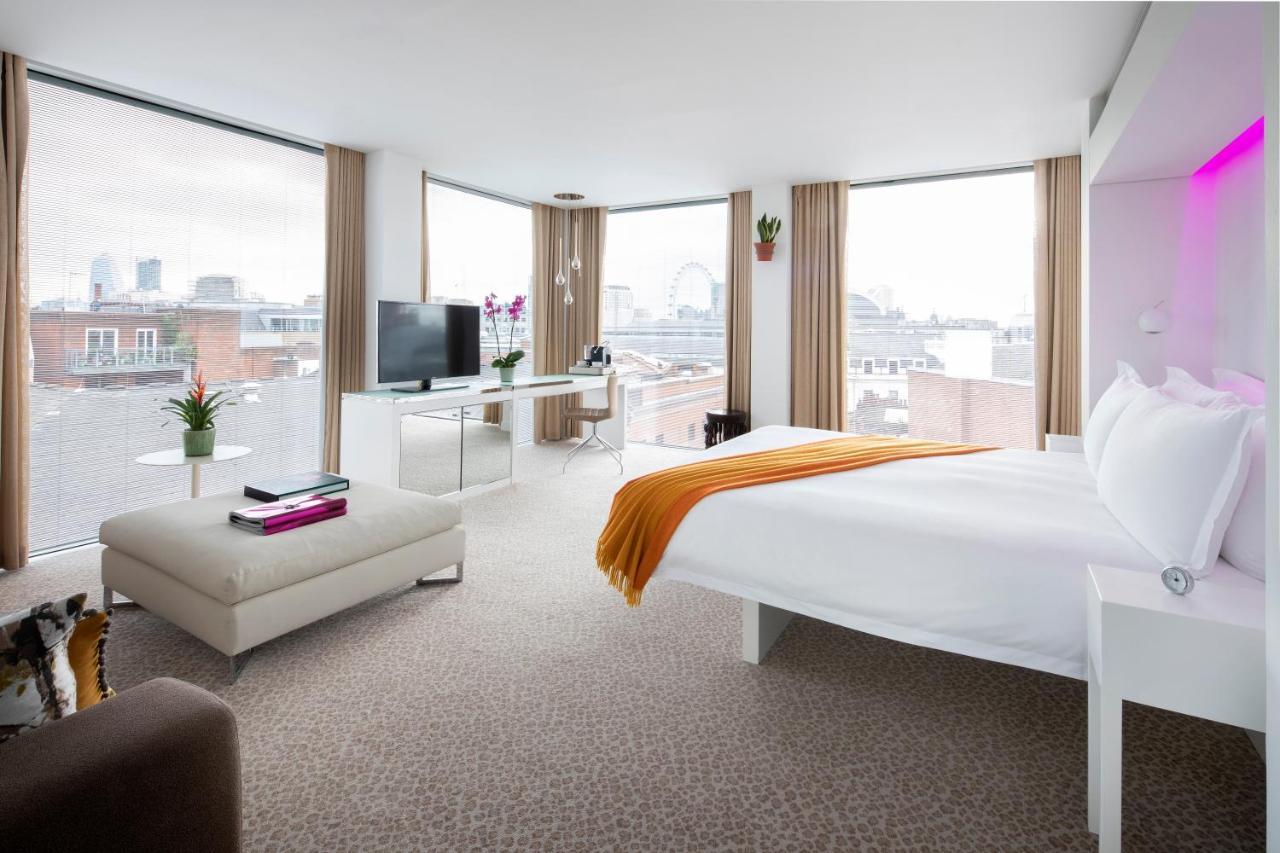 โรงแรมเซนต์ มาร์ติน เลน ลอนดอน ภายนอก รูปภาพ The photo shows a modern hotel room featuring a large bed with white linens and an orange throw blanket. The room has ample natural light coming through large windows with beige curtains, offering a view of a cityscape. There is a small seating area 