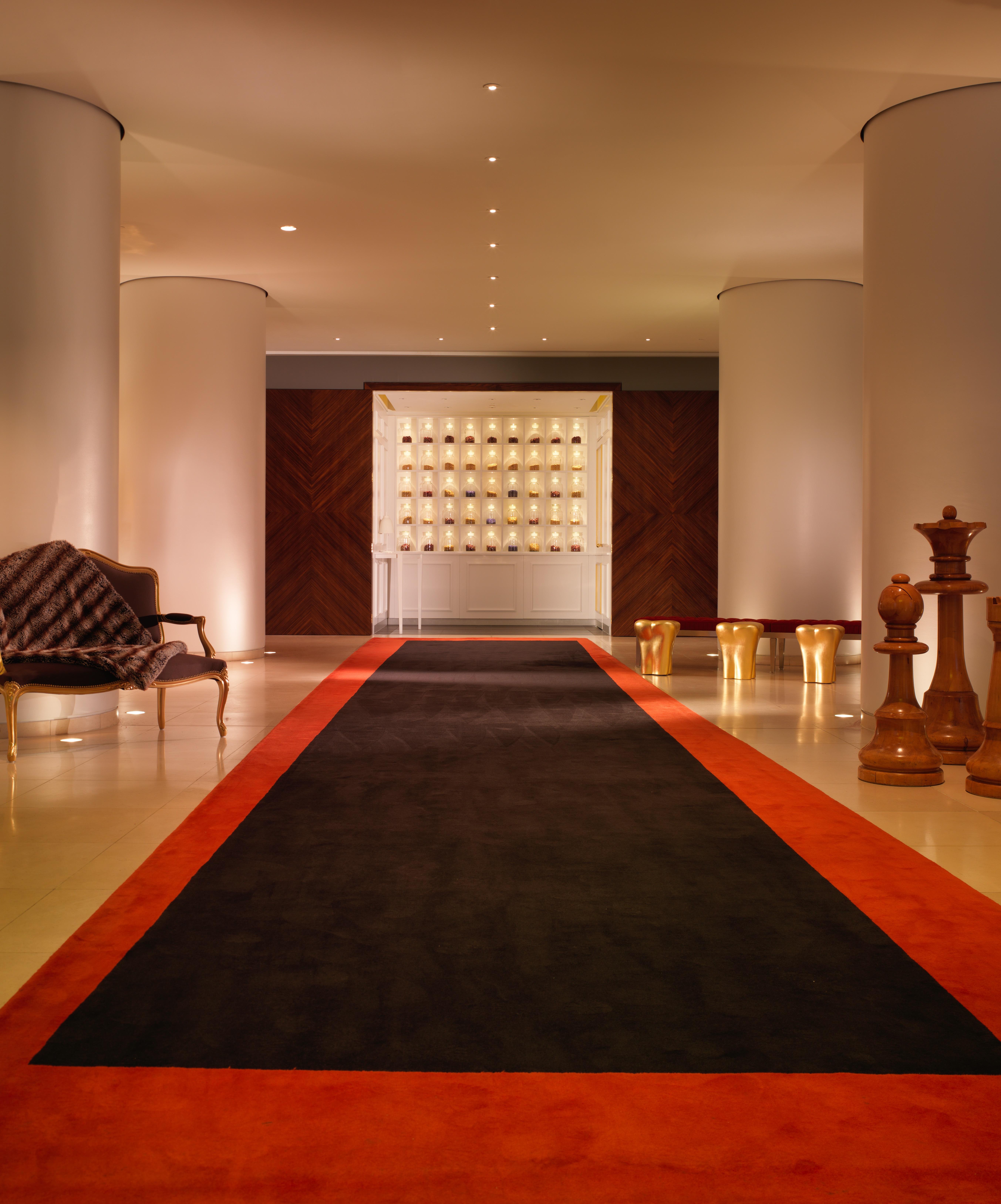 โรงแรมเซนต์ มาร์ติน เลน ลอนดอน ภายนอก รูปภาพ The photo shows a modern, elegant hallway or lobby area. There is a dark carpet runner in the center, bordered by a vibrant orange strip. On the left side, there is a stylish armchair, and on the right, large wooden pieces that resemble chess pieces.