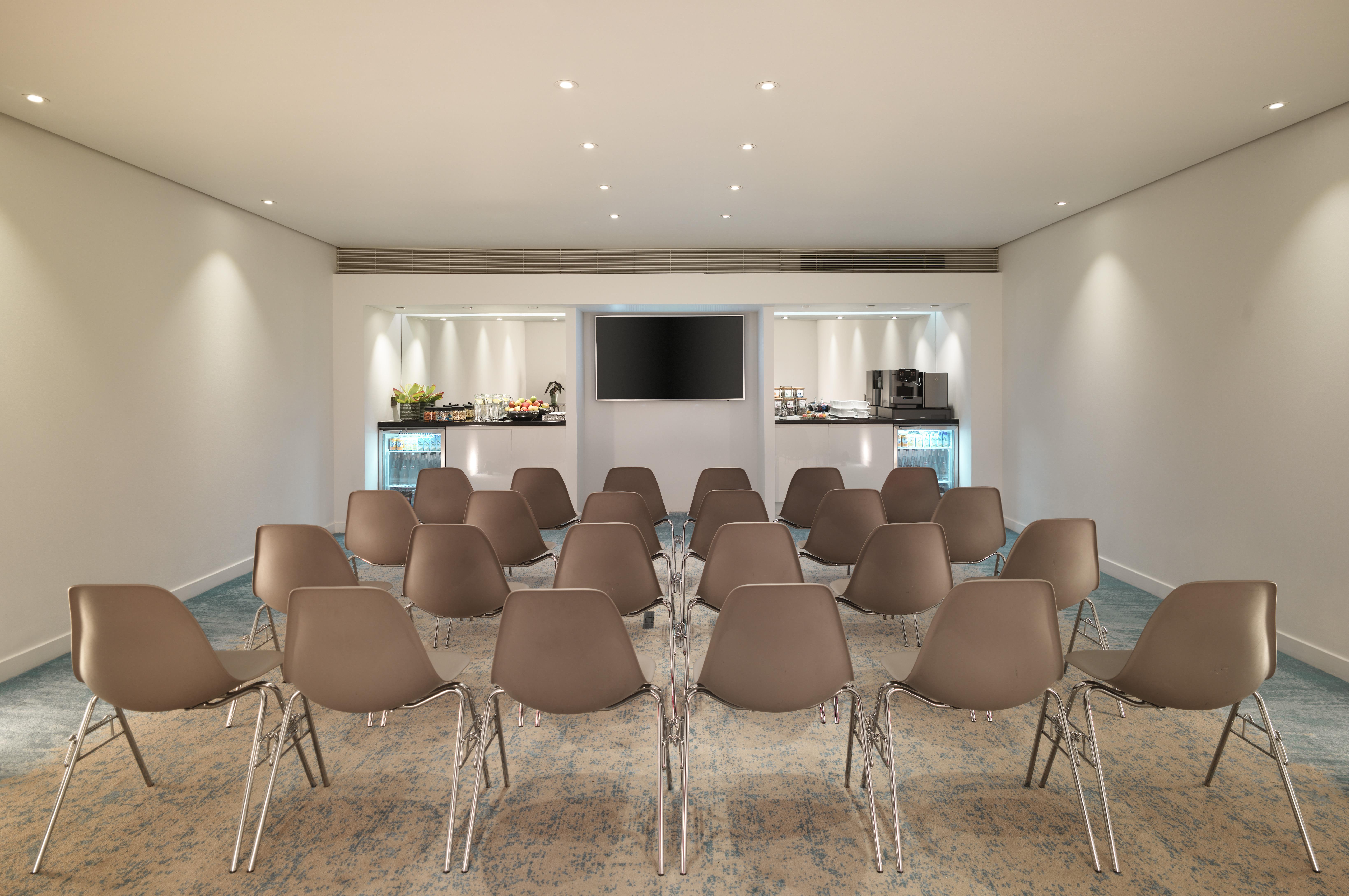 โรงแรมเซนต์ มาร์ติน เลน ลอนดอน ภายนอก รูปภาพ The photo depicts a modern meeting room or conference space. The room features several rows of light brown chairs arranged facing a wall-mounted television. In the background, there's a counter with refreshments, likely including food and beverages, 