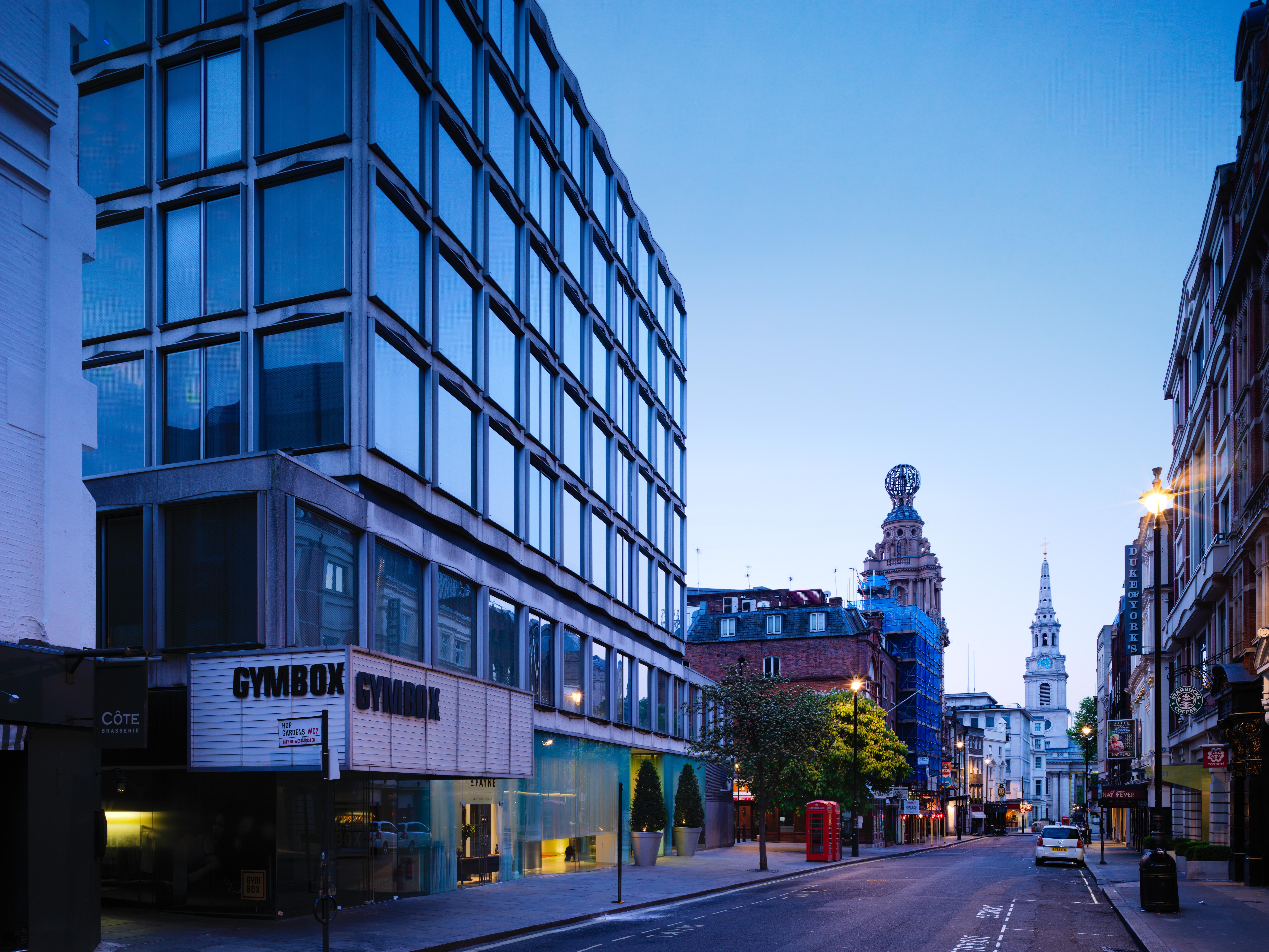 โรงแรมเซนต์ มาร์ติน เลน ลอนดอน ภายนอก รูปภาพ The photo shows a street scene during twilight, featuring a modern, glass-fronted building on the left side, which appears to be a gym called "GYMBOK GYM." On the right, there are more traditional architectural elements, including a tower and a steep