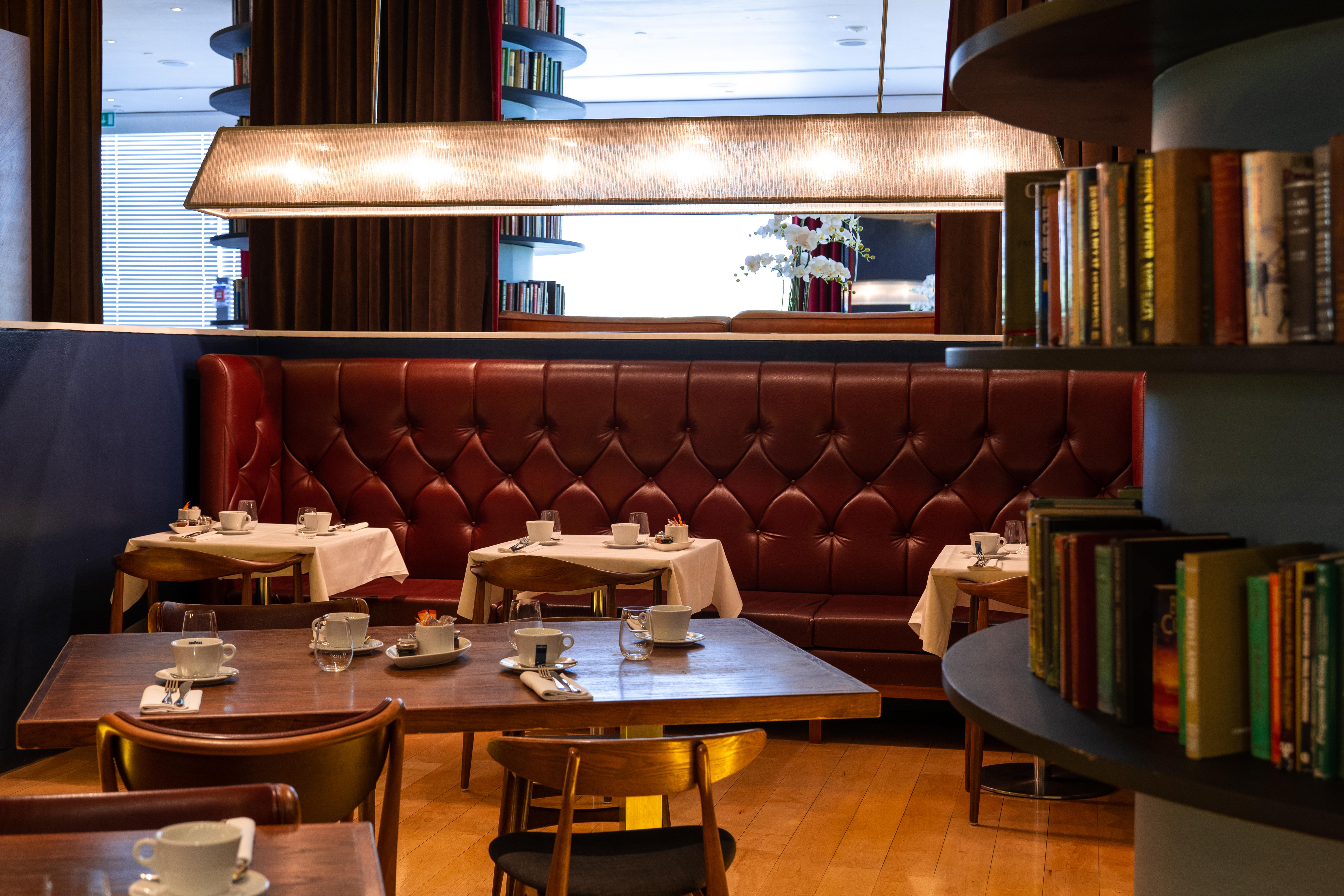 โรงแรมเซนต์ มาร์ติน เลน ลอนดอน ภายนอก รูปภาพ The photo depicts a cozy, elegantly designed restaurant interior. There are several round tables set with white tablecloths and accompanied by small white cups, likely for tea or coffee. The seating includes a plush, burgundy leather banquette agains
