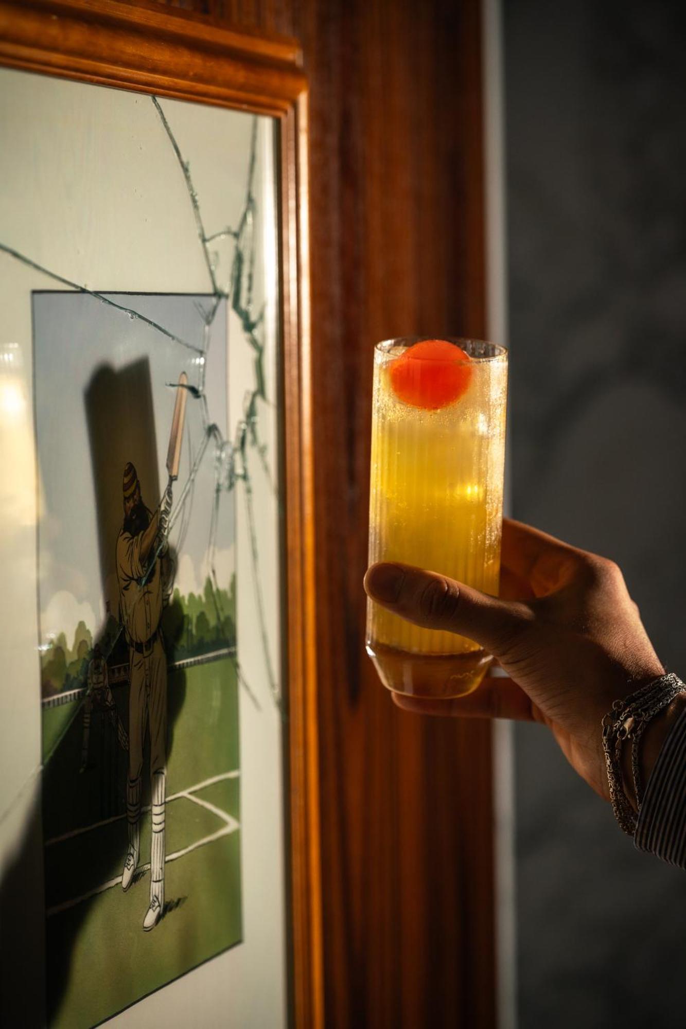 โรงแรมเซนต์ มาร์ติน เลน ลอนดอน ภายนอก รูปภาพ The photo shows a hand holding a tall glass containing a drink, which appears to be a yellow cocktail topped with a bright red cherry. In the background, there is a piece of art or a photograph that features a figure, possibly in a baseball or sporti
