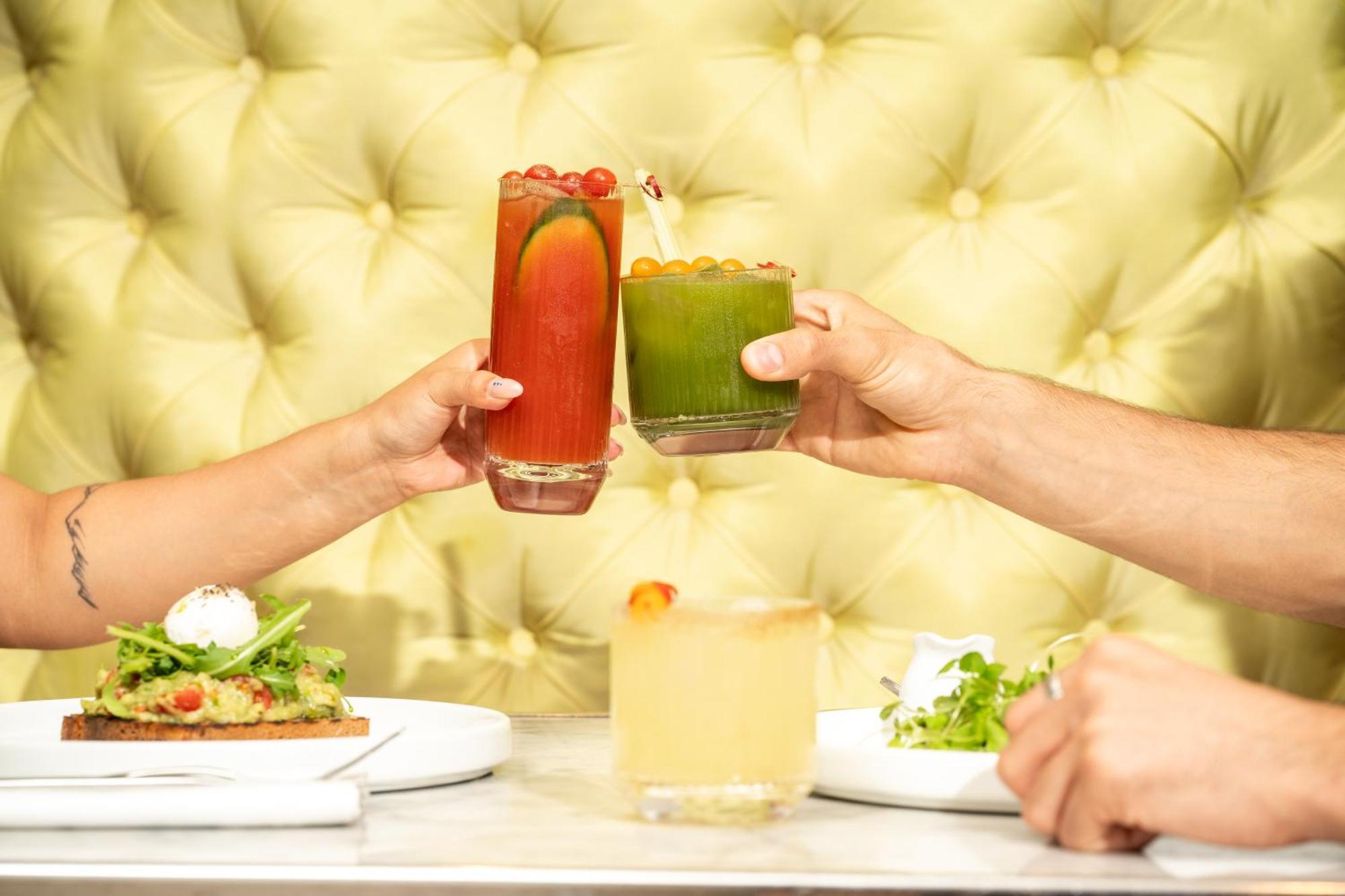 โรงแรมเซนต์ มาร์ติน เลน ลอนดอน ภายนอก รูปภาพ The photo features a vibrant dining scene with two hands clinking glasses in a toast. One glass contains a red drink, possibly a cocktail, adorned with garnishes, while the other is a green beverage, likely a smoothie or juice. In the foreground, the