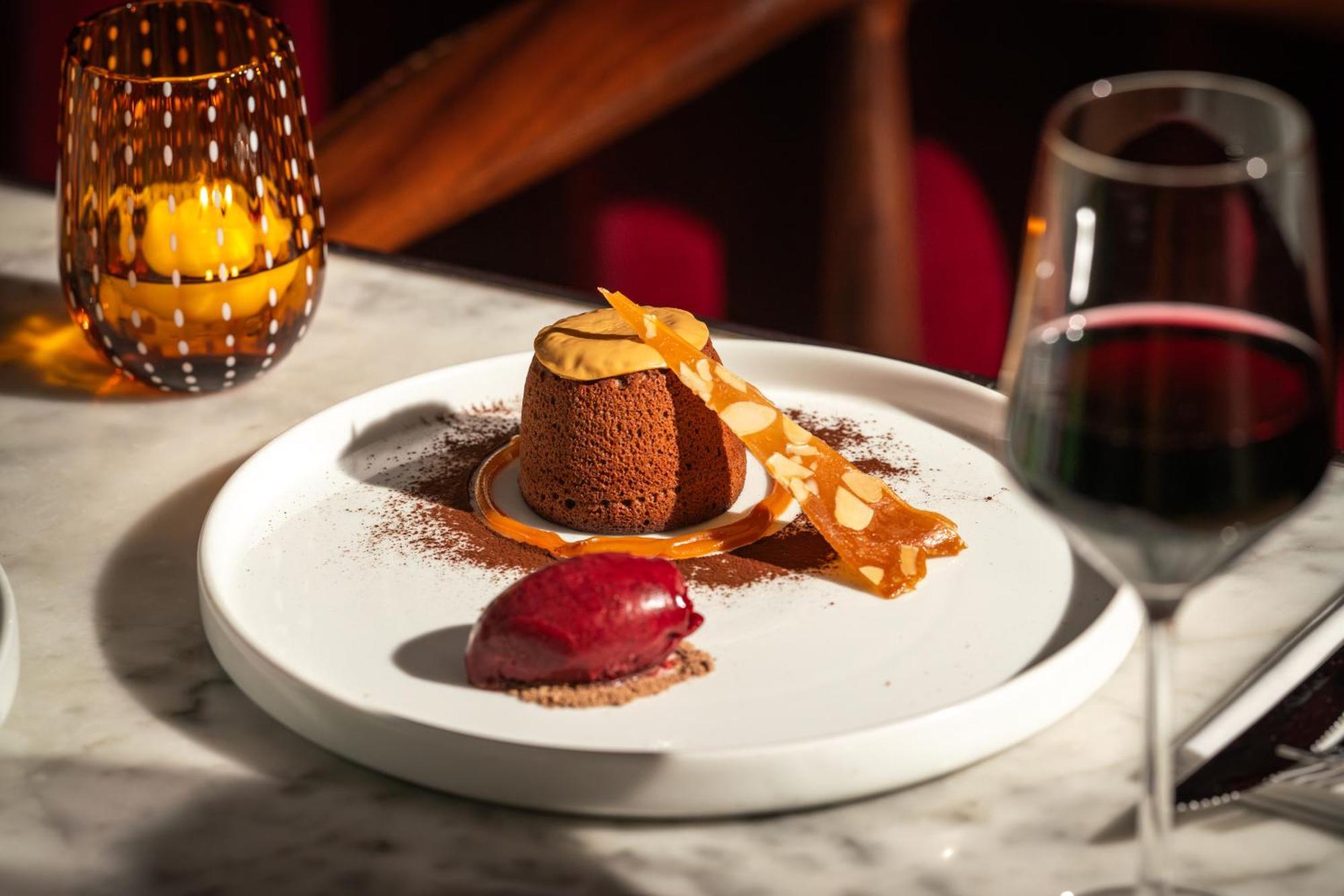 โรงแรมเซนต์ มาร์ติน เลน ลอนดอน ภายนอก รูปภาพ The photo showcases an elegantly presented dessert on a white plate. At the center is a chocolate dessert, likely a mousse or soufflé, adorned with a delicate strip of caramel or chocolate. Surrounding it is a sprinkle of cocoa powder. Beside the cho