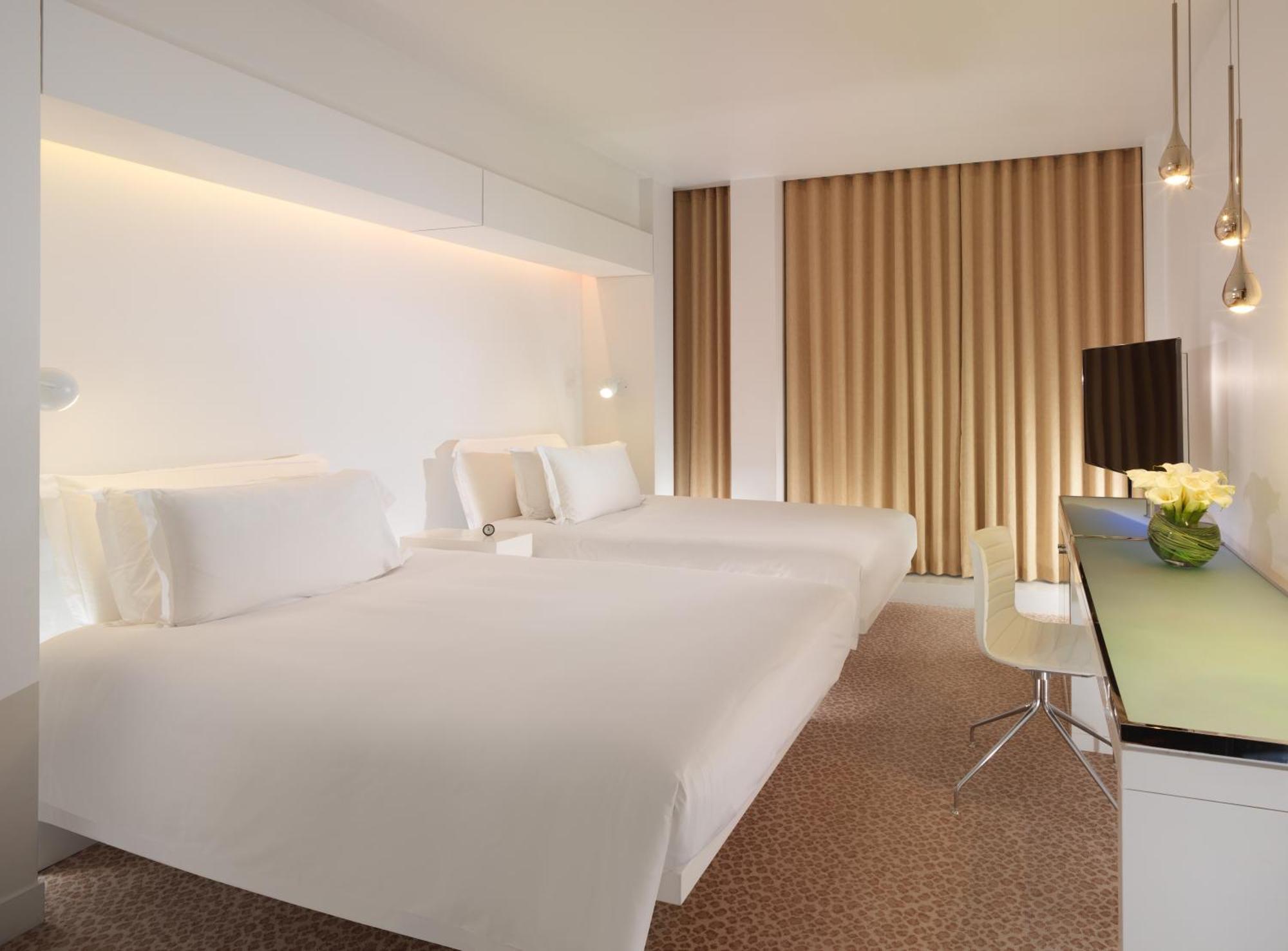 โรงแรมเซนต์ มาร์ติน เลน ลอนดอน ภายนอก รูปภาพ The photo shows a modern hotel room featuring two white beds neatly made with minimalistic design. The walls are primarily white with warm lighting highlighting the space. There are large windows dressed with light brown curtains. A small desk with a
