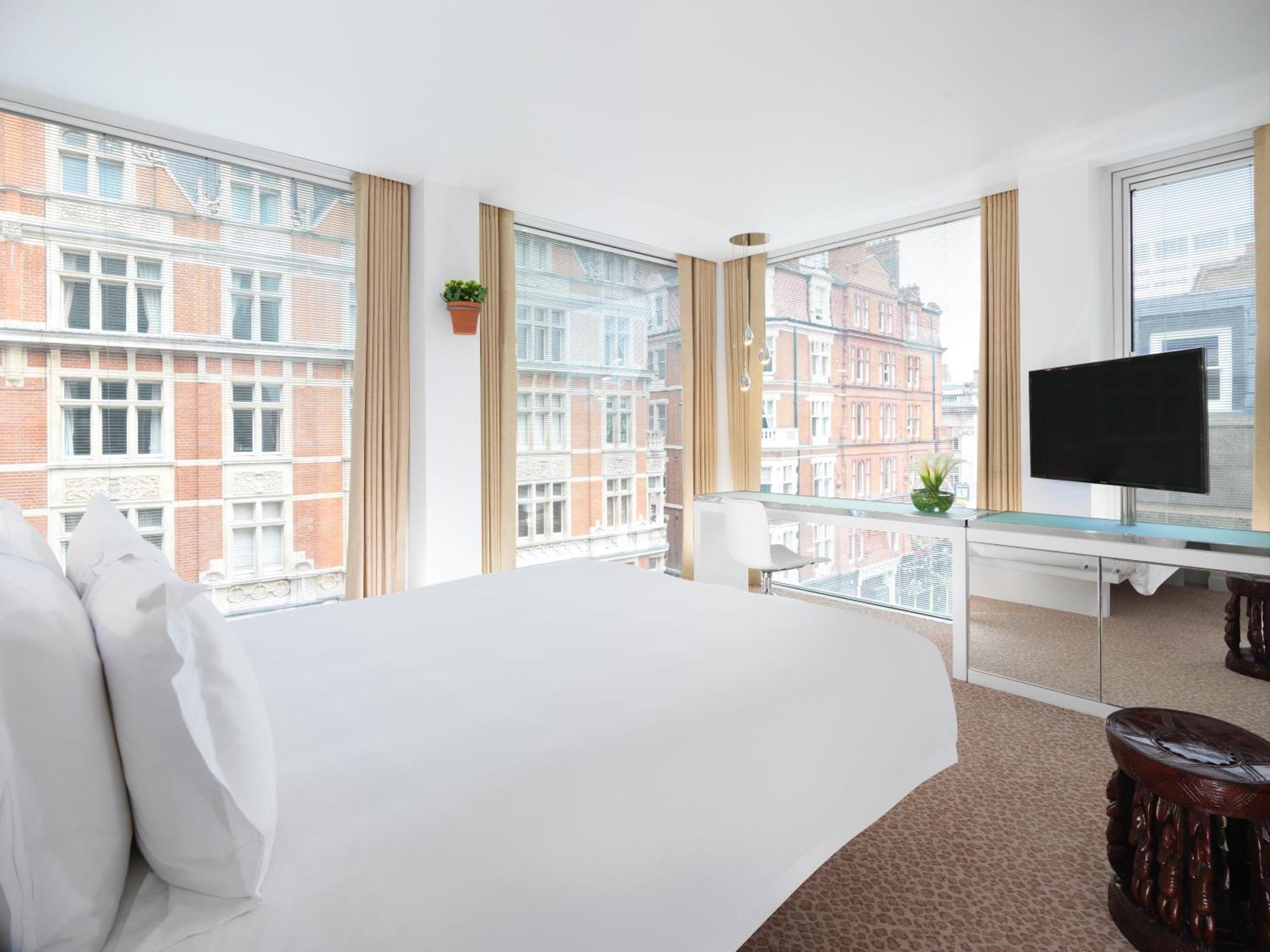 โรงแรมเซนต์ มาร์ติน เลน ลอนดอน ภายนอก รูปภาพ The photo shows a modern hotel room featuring a large bed with white linens, positioned near large windows that provide abundant natural light. The windows offer a view of brick buildings outside. There is a flat-screen TV mounted on the wall, a mini